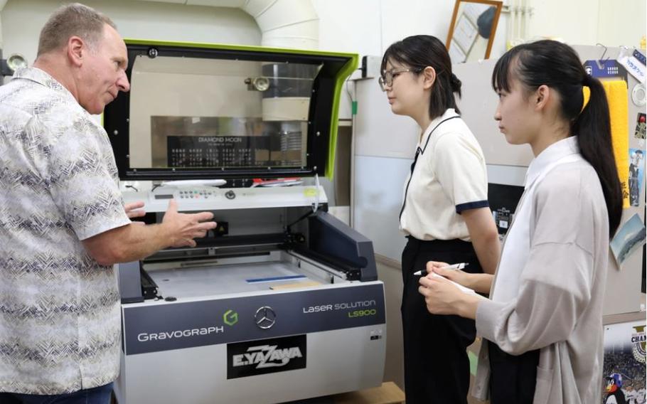 James McGee, manager of the Visual Information Division at U.S. Army Garrison Japan, shows Chihiro Suzuki, right, a sophomore at Toyoeiwa University and an intern at the USAG Japan Public Affairs Office and another intern VID’s engraving machine Aug 7.